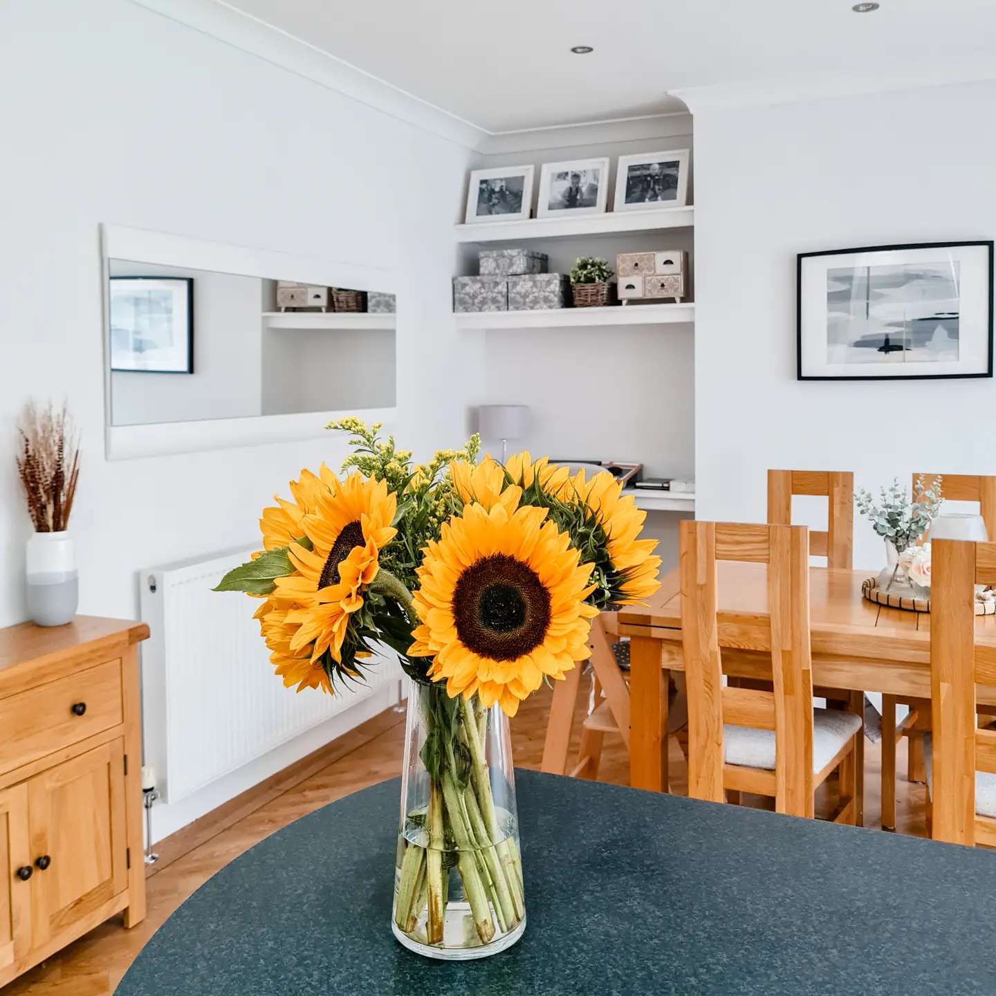 A dining table-dining room furniture-oak dining table and chairs-kitchen island-vase of sunflowers-white walls-long horizontal mirror-framed photography-oak sideboard-parquet flooring