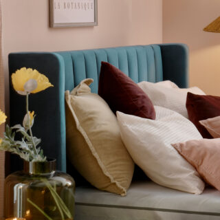 A bed and bedside table-bedroom furniture-teal upholstered bed-layered cushions-plain mattress-pink walls-green vase with flowers-candle light-cream and wood bedside table-floral wall art