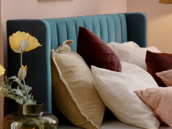 A bed and bedside table-bedroom furniture-teal upholstered bed-layered cushions-plain mattress-pink walls-green vase with flowers-candle light-cream and wood bedside table-floral wall art