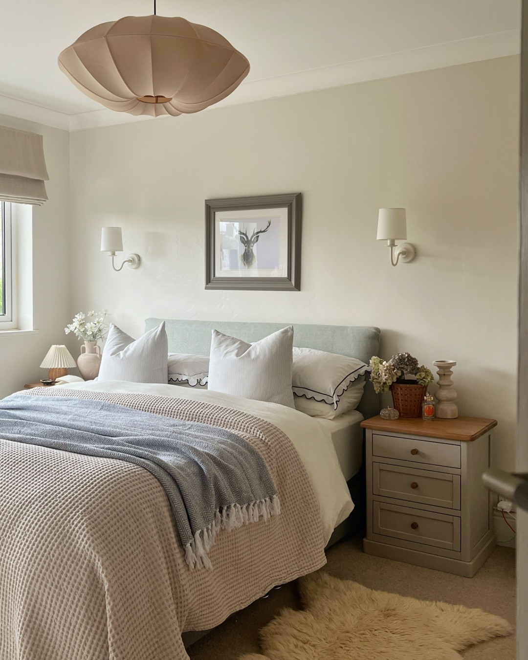 A bed and bedside table-bedroom furniture-neutral walls-upholstered bed-layered bedding-painted wooden bedside table-sheepskin rug-statement ceiling light-delicate wall lights-fresh flowers-natural light