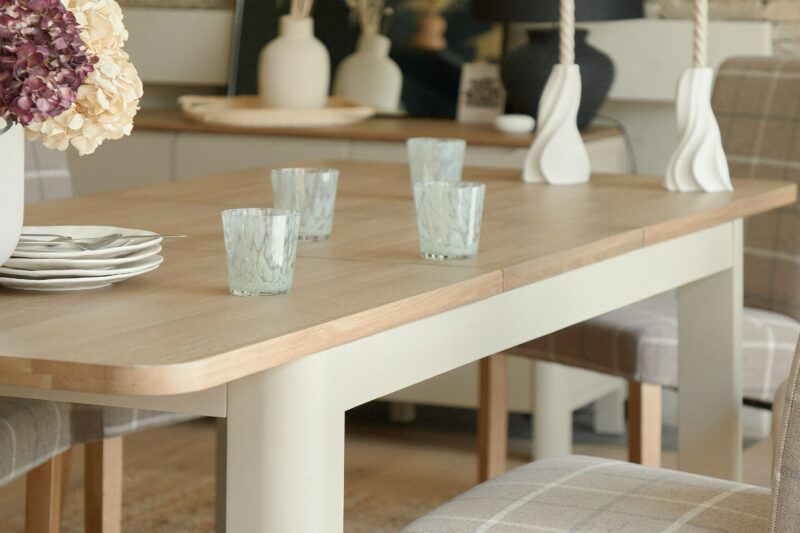 A dining table and chairs-dining room furniture-oak wooden tabletop-painted wooden legs-plaid upholstered chairs-mouth blown water glasses-sculptural candleholders-stacked white plates-vase of hydrangeas