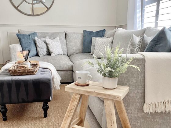 A modular corner sofa-living room furniture-pale grey corner sofa-denim blue accent cushions-dark grey footstool-rustic pale wooden stool-jute rug-cream walls-wooden floor-natural light