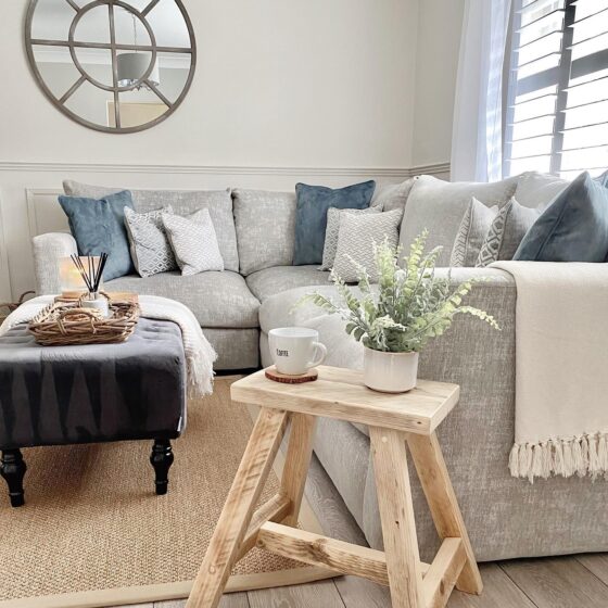 A modular corner sofa-living room furniture-pale grey corner sofa-denim blue accent cushions-dark grey footstool-rustic pale wooden stool-jute rug-cream walls-wooden floor-natural light