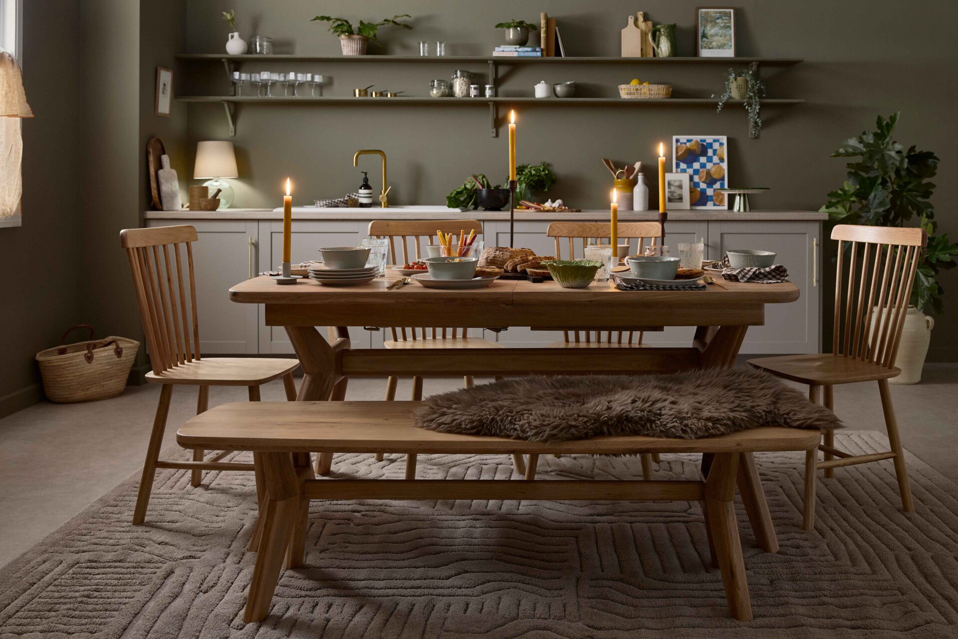 A dining table, display cabinet and bench-kitchen diner furniture-oak dining table set-neutral textured rug-spindle back chairs-olive green walls-sheepskin rug-tapered candles-soup bowls-bistro curtain-grey kitchen-open kitchen shelving-brass tap-autumnal tablescape