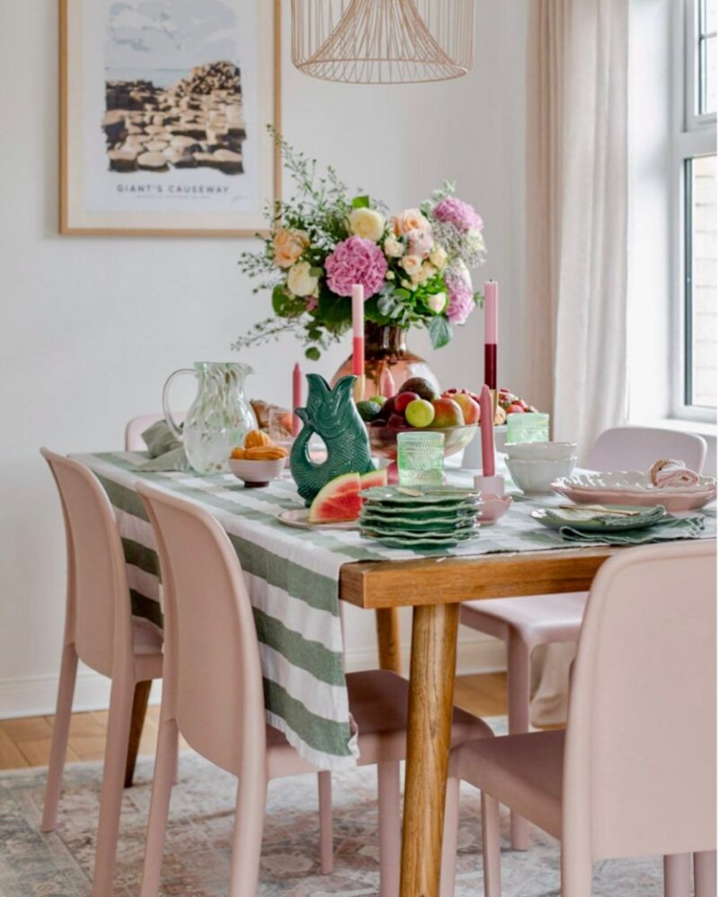 A dining table and chairs-dining room furniture-pink dining chairs-oak dining table-striped tablecloth-stacked crockery-tapered candles-green fish jug-fresh fruit bowl-landscape wall art-metal painted ceiling light-natural light-draped sheer curtain-vintage style rug