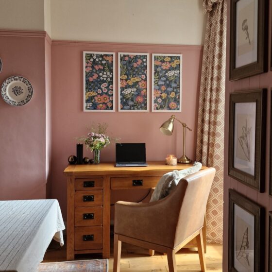 A desk and chair-home office furniture-oak desk with drawers-brown leather look upholstered chair-pink and neutral walls-framed floral wallpaper-botanical artwork-decorative wall plates-patterned curtain-vintage rug-beige tablecloth