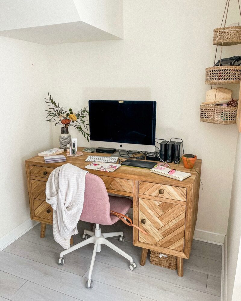 A desk and chair-home office furniture-oak desk with drawers-pink upholstered office chair-cream walls-grey wooden flooring-desktop computer-vase of flowers-notebooks-hanging rattan storage