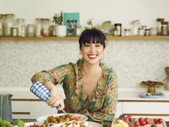 A woman in a kitchen-cooking a mixture of sharing dished-pouring oil-blue and white check olive oil bottle-fresh herbs-chopping fruit and vegetables-open shelf-kitchen units-floral patterned dress