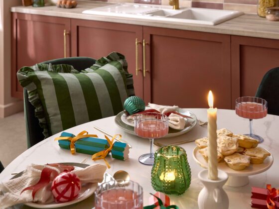 A dining table and chairs-kitchen diner furniture-white ceramic tabletop-green velvet dining chairs-stripey green cushion with frills-christmas tablescape-festive place settings-colourful baubles-striped crackers-twisted candles-green glass tealight-scalloped champagne glasses-burgundy kitchen units-brass door handles