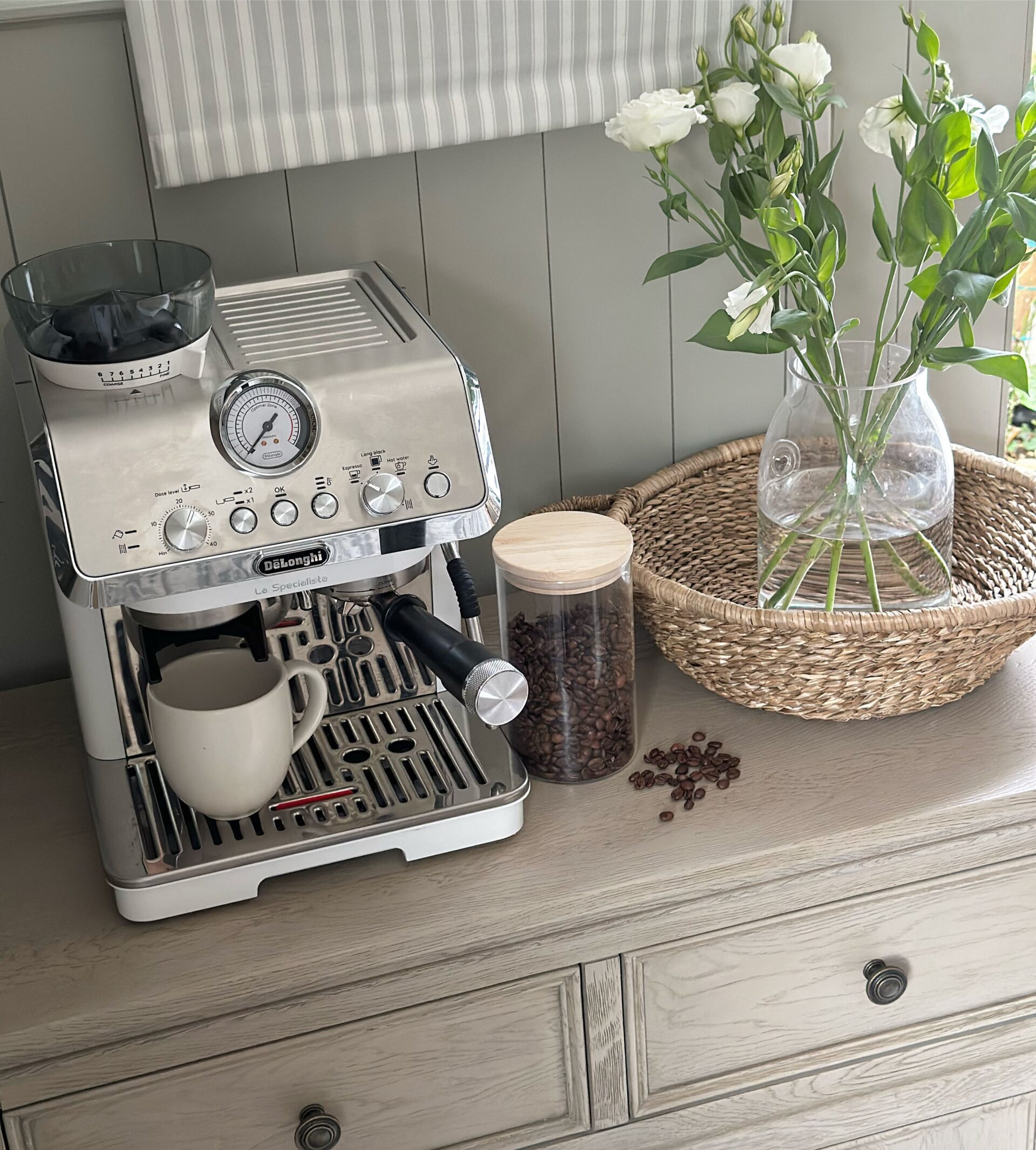A sideboard-dining room furniture-coffee station-coffee machine-coffee beans in cannister-woven tray-fresh flowers in glass vase-weathered oak sideboard-neutral walls with panelling-striped blind
