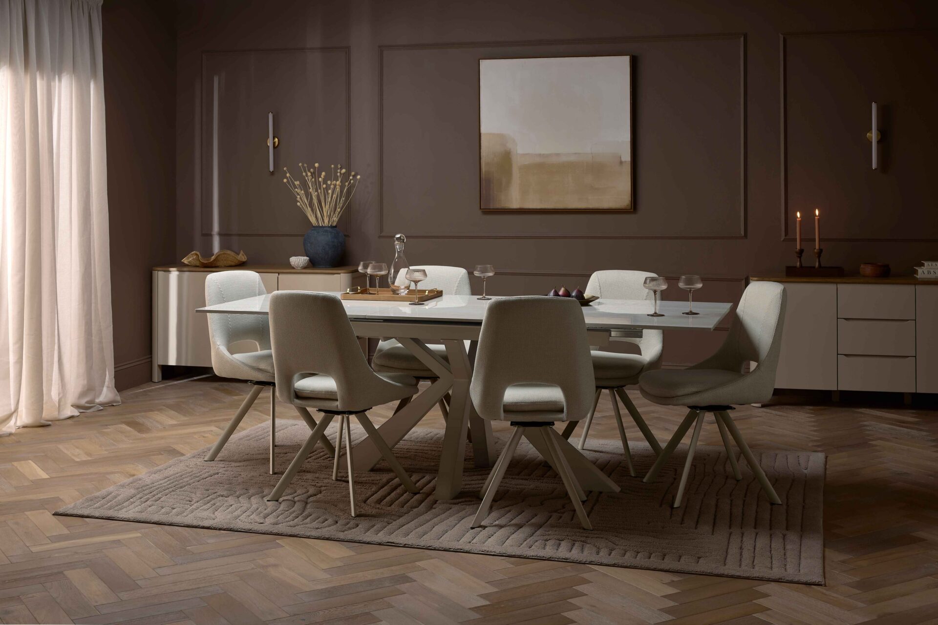 A dining table and chairs-dining room furniture-neutral fabric dining chairs-ceramic dining table-parquet floor-textured neutral rug-dark brown walls-painted cream sideboards-abstract wall art-tapered candles-dried flowers-wall lights-sheer draped curtains