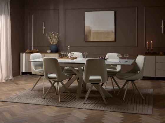 A dining table and chairs-dining room furniture-neutral fabric dining chairs-ceramic dining table-parquet floor-textured neutral rug-dark brown walls-painted cream sideboards-abstract wall art-tapered candles-dried flowers-wall lights-sheer draped curtains