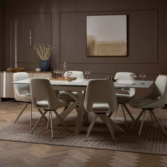 A dining table and chairs-dining room furniture-neutral fabric dining chairs-ceramic dining table-parquet floor-textured neutral rug-dark brown walls-painted cream sideboards-abstract wall art-tapered candles-dried flowers-wall lights-sheer draped curtains