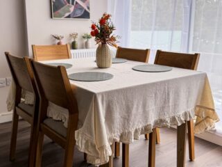 A dining table and chairs-dining room furniture-neutral frilly tablecloth-neutral walls-sheer curtains-floral wall art-wooden and fabric chairs-round placemats-vase of flowers-wooden floor