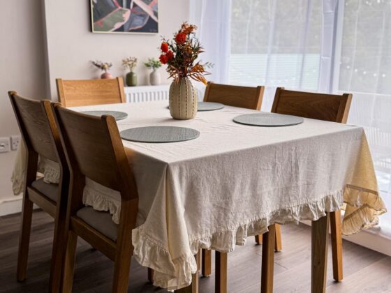 A dining table and chairs-dining room furniture-neutral frilly tablecloth-neutral walls-sheer curtains-floral wall art-wooden and fabric chairs-round placemats-vase of flowers-wooden floor