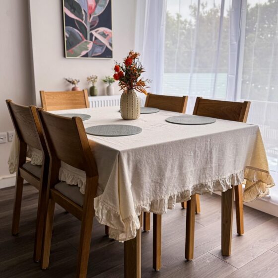A dining table and chairs-dining room furniture-neutral frilly tablecloth-neutral walls-sheer curtains-floral wall art-wooden and fabric chairs-round placemats-vase of flowers-wooden floor