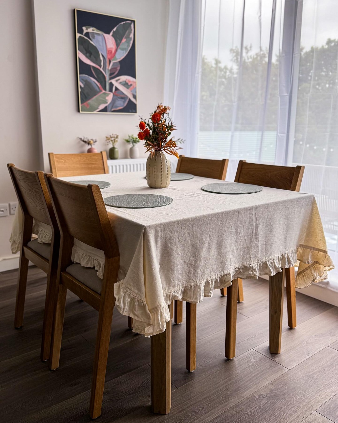 A dining table and chairs-dining room furniture-neutral frilly tablecloth-neutral walls-sheer curtains-floral wall art-wooden and fabric chairs-round placemats-vase of flowers-wooden floor