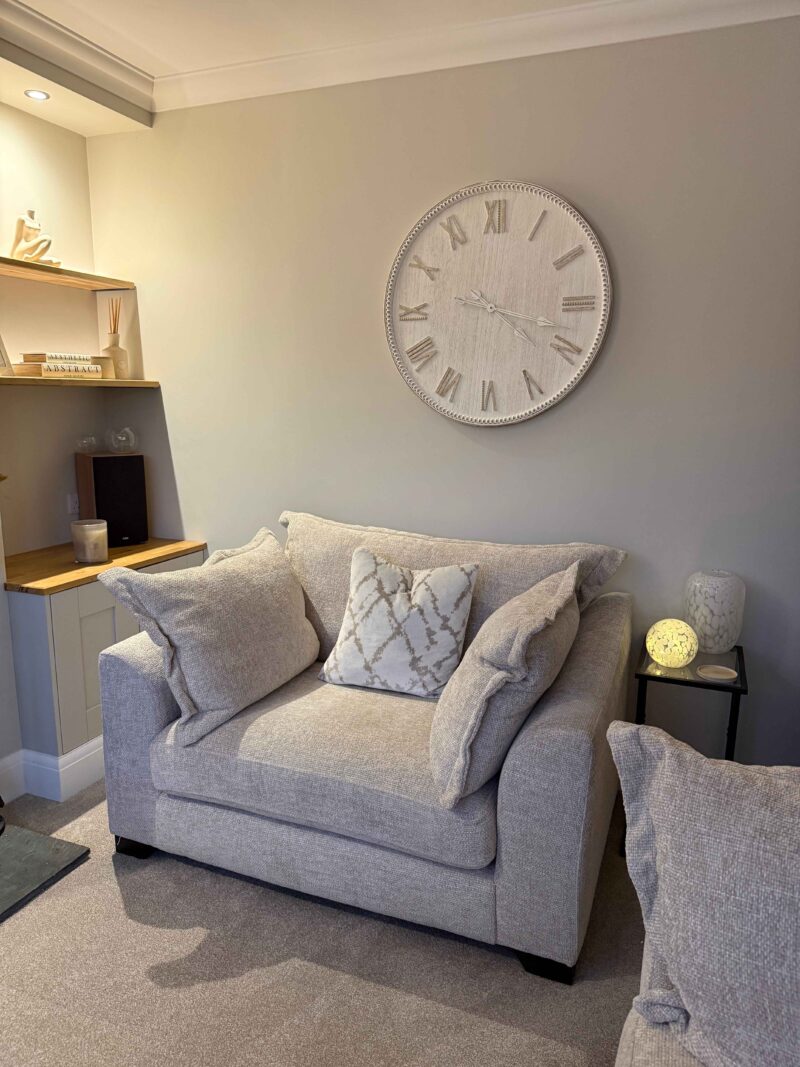A loveseat-living room furniture-beige loveseat-neutral armchair-textured scatter cushions-beige carpets-neutral walls-oversized wall clock-built in shelving-metal and glass side table-glass orb table light