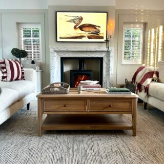 A coffee table and sofas-wooden coffee table with storage-textured neutral rug-marble fireplace-black log burner-cream sofas-striped red and cream cushion and throw-neutral walls-stacked books-rattan tray-art TV-window shutters