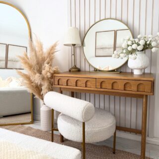 A dressing table-bedroom furniture-wooden console table with arch detail-boucle dressing table chair-round brass mirror-table lamp-vase of white flowers-vase of pampas grass-fluted wall panelling-white walls-floor standing arched mirror-jute rug