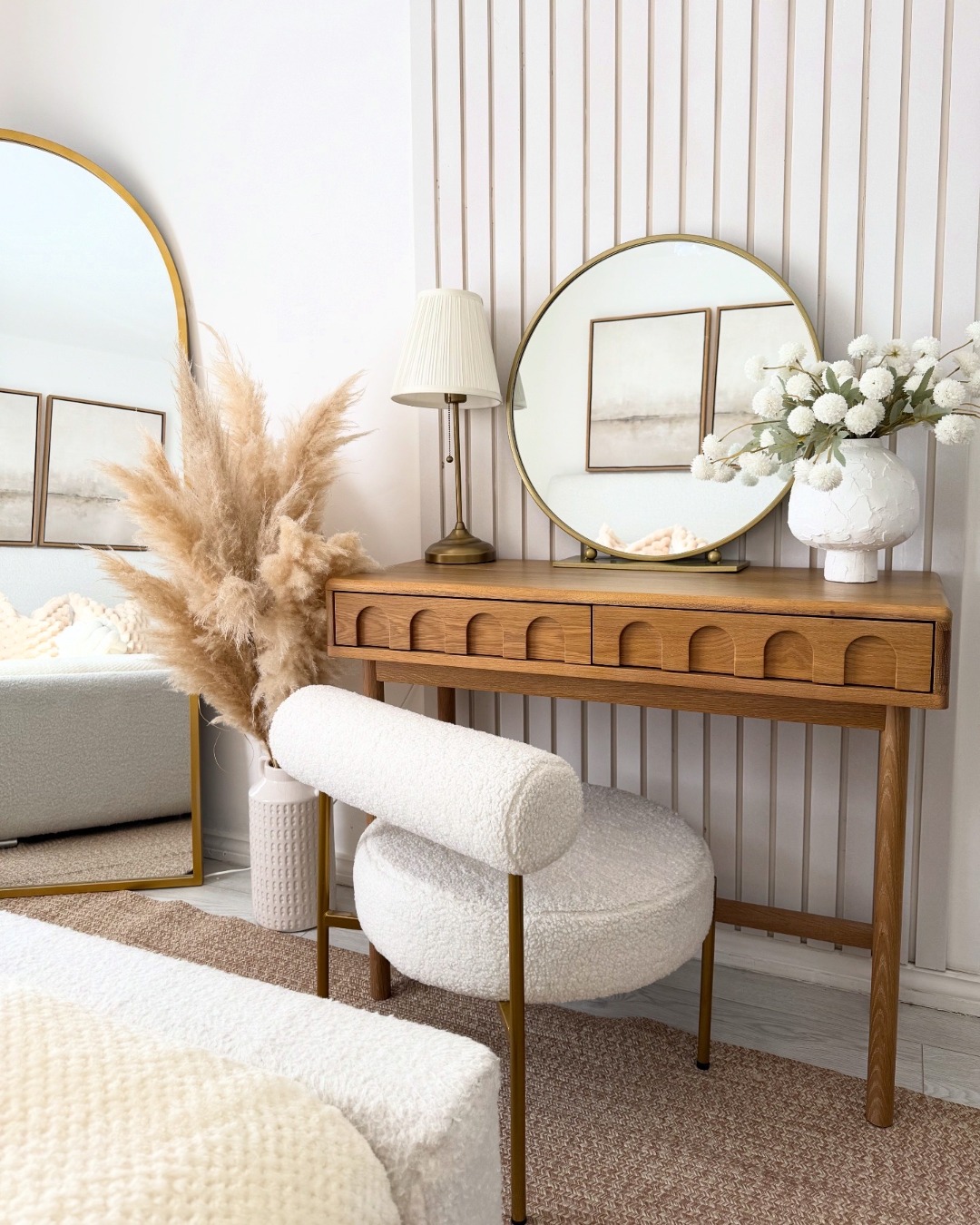 A dressing table-bedroom furniture-wooden console table with arch detail-boucle dressing table chair-round brass mirror-table lamp-vase of white flowers-vase of pampas grass-fluted wall panelling-white walls-floor standing arched mirror-jute rug