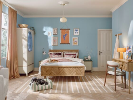 A bed, wardrobe, and dressing table-bedroom furniture-wooden double bed-neutral painted double wardrobe-sky blue walls-striped natural rug-wooden dressing table-dark wood upholstered mid century style chair-white bedroom door-art wall-striped dog bed-grey dog-parquet wooden floor-draped curtains-cream armchair