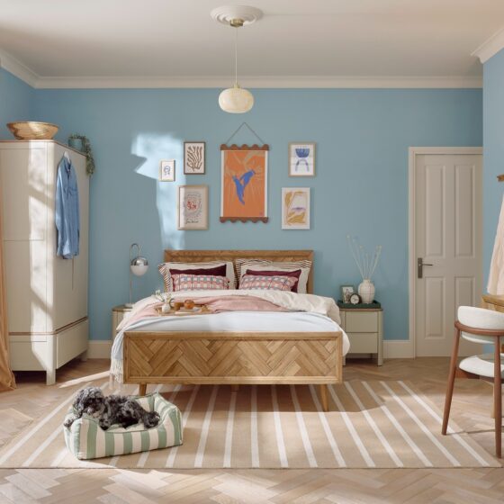 A bed, wardrobe, and dressing table-bedroom furniture-wooden double bed-neutral painted double wardrobe-sky blue walls-striped natural rug-wooden dressing table-dark wood upholstered mid century style chair-white bedroom door-art wall-striped dog bed-grey dog-parquet wooden floor-draped curtains-cream armchair