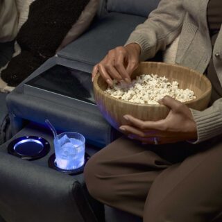 A sofa-living room furniture-dark blue recliner sofa with console screen and cup holders-tactile cushion-armrest-iced drink in glass cup with straw-person eating popcorn-wooden bowl-neutral clothing