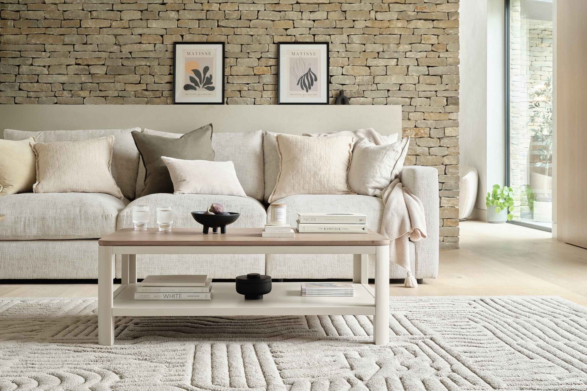 A sofa, coffee table, and rug-living room furniture-beige sofa-neutral cushions-neutral textured rug-wooden and painted cream coffee table with shelf-exposed brick wall-framed wall art-sculptural objects-window wall-neutral wall-potted house plant