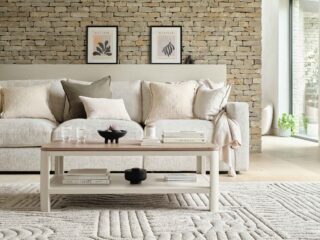 A sofa, coffee table, and rug-living room furniture-beige sofa-neutral cushions-neutral textured rug-wooden and painted cream coffee table with shelf-exposed brick wall-framed wall art-sculptural objects-window wall-neutral wall-potted house plant