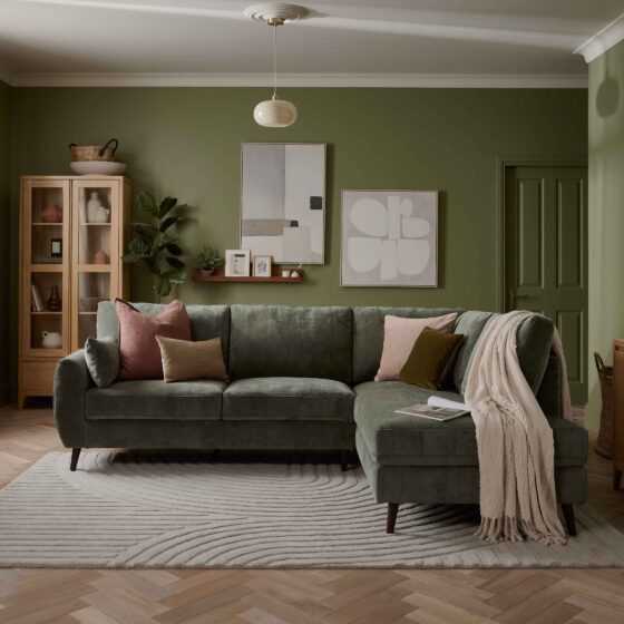 A sofa, sideboard, and display cabinet-living room furniture-green corner sofa-green walls-neutral ceiling-glass ceiling light-wooden sideboard-wood and glass display cabinet-neutral abstract wall art-floating wooden shelf-sheer draped curtains-textured neutral rug-wooden parquet floor-draped beige throw-scatter cushions
