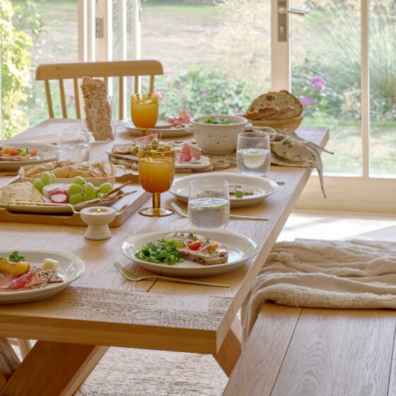 A dining table-dining room furniture-an oak dining table-oak bench-oak spindle back chairs-amber glassware-white tableware-gold cutlery-water glasses-food serving platter-salad-bread basket-wooden flooring-beige throw-jute rug-french doors-garden view