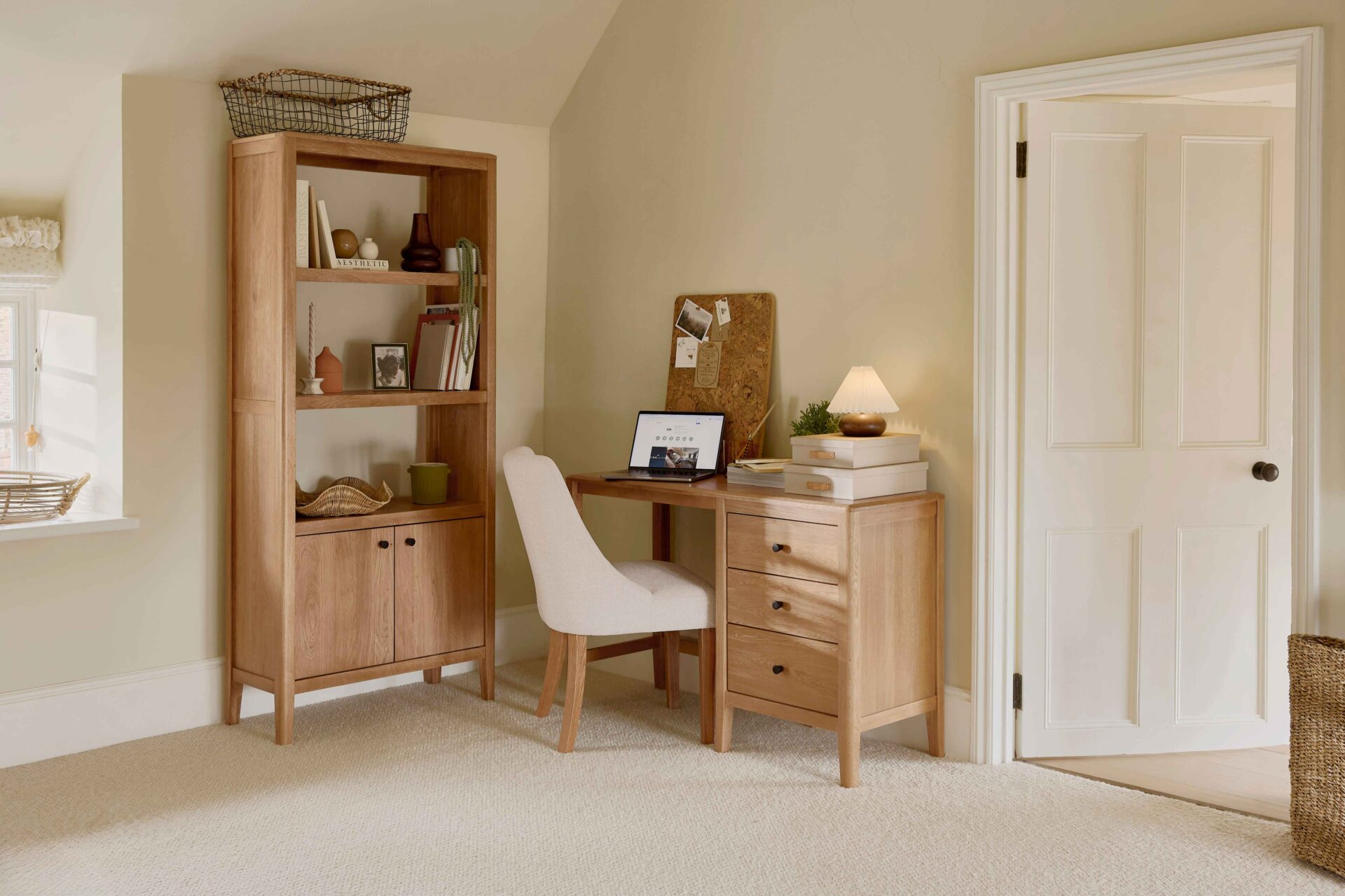 A desk, chair, and bookcase-home office furniture-natural oak open shelving-natural oak desk with drawer storage-cream upholstered office chair-woven storage basket-stacked storage boxes-cork board-laptop-pleated table lamp-beige walls-off white woodwork-beige carpet-books-potted houseplants-vases