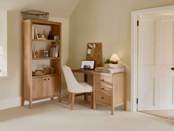 A desk, chair, and bookcase-home office furniture-natural oak open shelving-natural oak desk with drawer storage-cream upholstered office chair-woven storage basket-stacked storage boxes-cork board-laptop-pleated table lamp-beige walls-off white woodwork-beige carpet-books-potted houseplants-vases