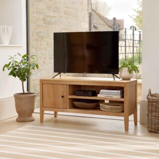 A TV unit-living room furniture-oak TV unit-striped jute rug-woven storage basket-faux brown sheepskin rug-clay pot with houseplant-floor standing rattan and brass lamp-marble fireplace-window wall-off white walls-white vase-stacked books-woven tray-TV