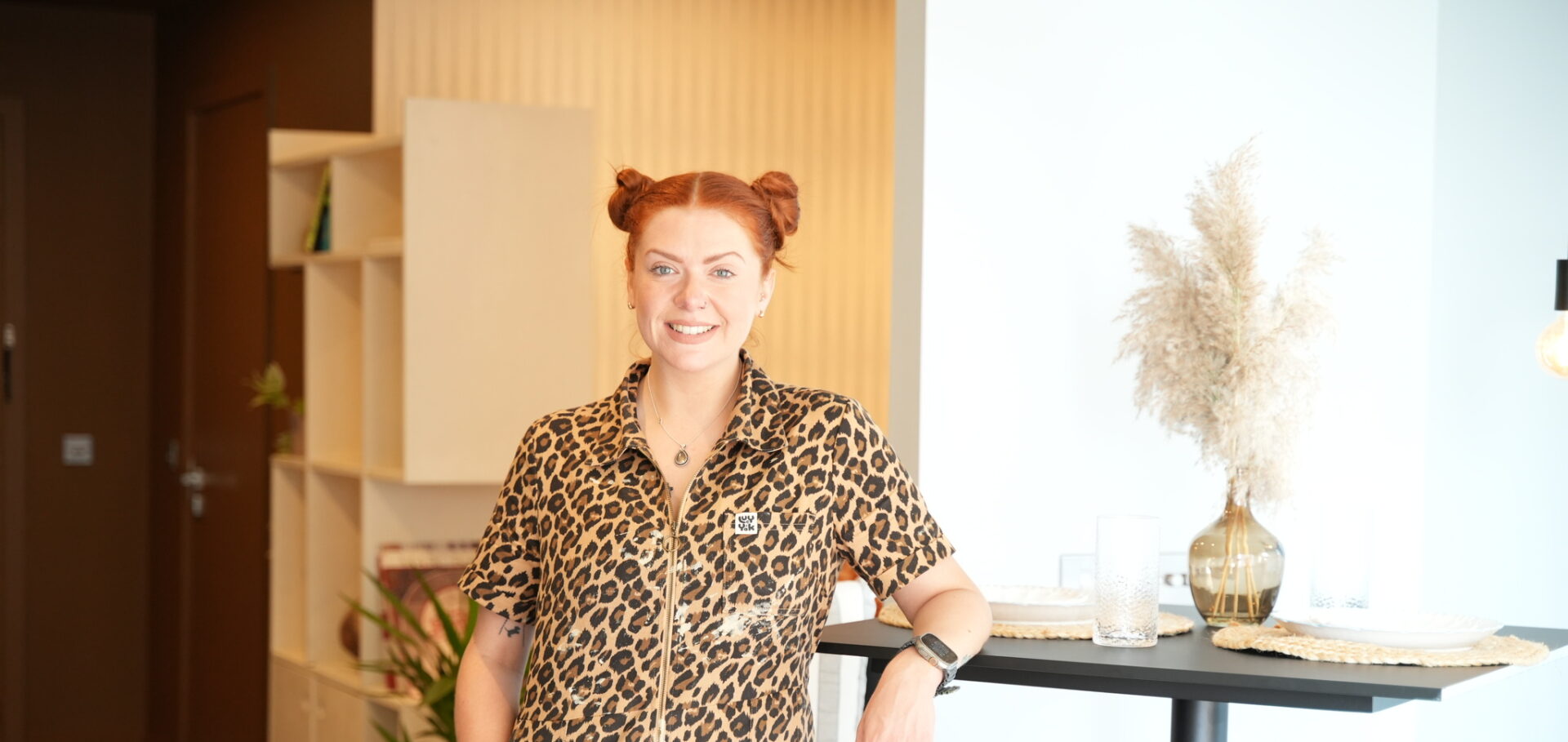 A woman and breakfast bar table-open plan living furniture-woman in leopard print overalls-red hair in bunches-black breakfast bar table-black stools-vase with pampas grass-soft blue walls-neutral walls-brown walls-built in open shelving-wooden floor-potted houseplant