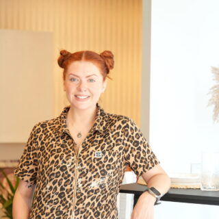 A woman and breakfast bar table-open plan living furniture-woman in leopard print overalls-red hair in bunches-black breakfast bar table-black stools-vase with pampas grass-soft blue walls-neutral walls-brown walls-built in open shelving-wooden floor-potted houseplant