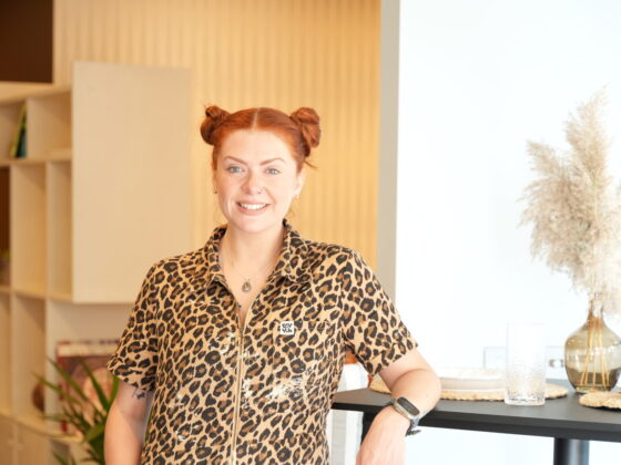 A woman and breakfast bar table-open plan living furniture-woman in leopard print overalls-red hair in bunches-black breakfast bar table-black stools-vase with pampas grass-soft blue walls-neutral walls-brown walls-built in open shelving-wooden floor-potted houseplant