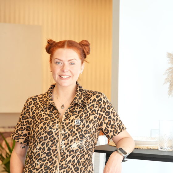 A woman and breakfast bar table-open plan living furniture-woman in leopard print overalls-red hair in bunches-black breakfast bar table-black stools-vase with pampas grass-soft blue walls-neutral walls-brown walls-built in open shelving-wooden floor-potted houseplant
