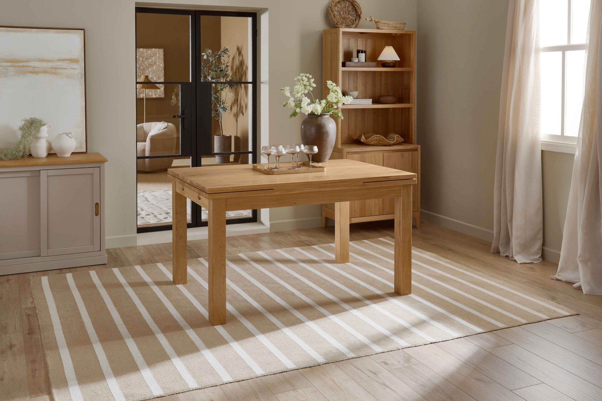 A wooden dining table and small dresser-dining room furniture-natural oak wooden dining table-neutral walls-natural oak small wooden dresser-draped neutral curtains-crittall internal door-striped jute rug-wooden floor-stone painted sideboard-champagne glasses-abstract neutral artwork-brown ceramic vase-tray