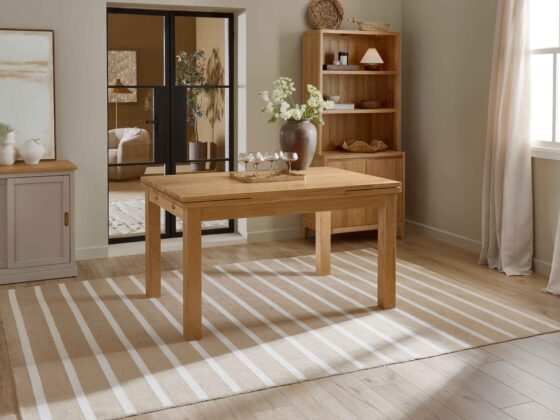 A wooden dining table and small dresser-dining room furniture-natural oak wooden dining table-neutral walls-natural oak small wooden dresser-draped neutral curtains-crittall internal door-striped jute rug-wooden floor-stone painted sideboard-champagne glasses-abstract neutral artwork-brown ceramic vase-tray