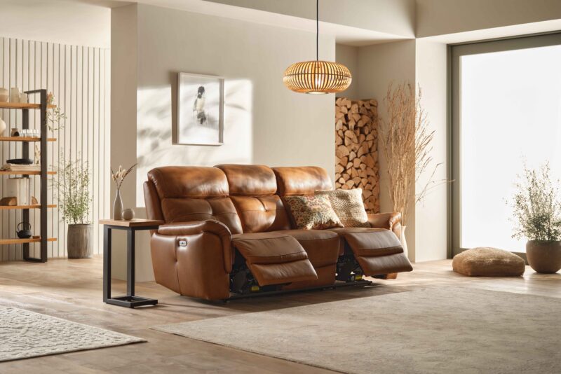 A leather sofa-living room furniture-brown leather recliner sofa-textured cream cushions-stacked wood pile-rattan ceiling light-wooden floor-neutrals rugs-window wall-floor cushion-potted houseplant-wood and metal side table-painted wooden panelling-neutral walls-wood and metal open shelving unit-framed print