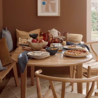 A dining table, chairs, and benches-dining room furniture-natural oak oval dining table-wishbone style dining chairs-natural oak benches-warm brown walls-abstract wall art-autumnal tablescape-neutral textured rug-sisal carpet-woven storage basket-ceramic fruit bowl filled with apples-baked apple tart-off white french doors-amber water jug-tea pot-crockery-scatter cushions