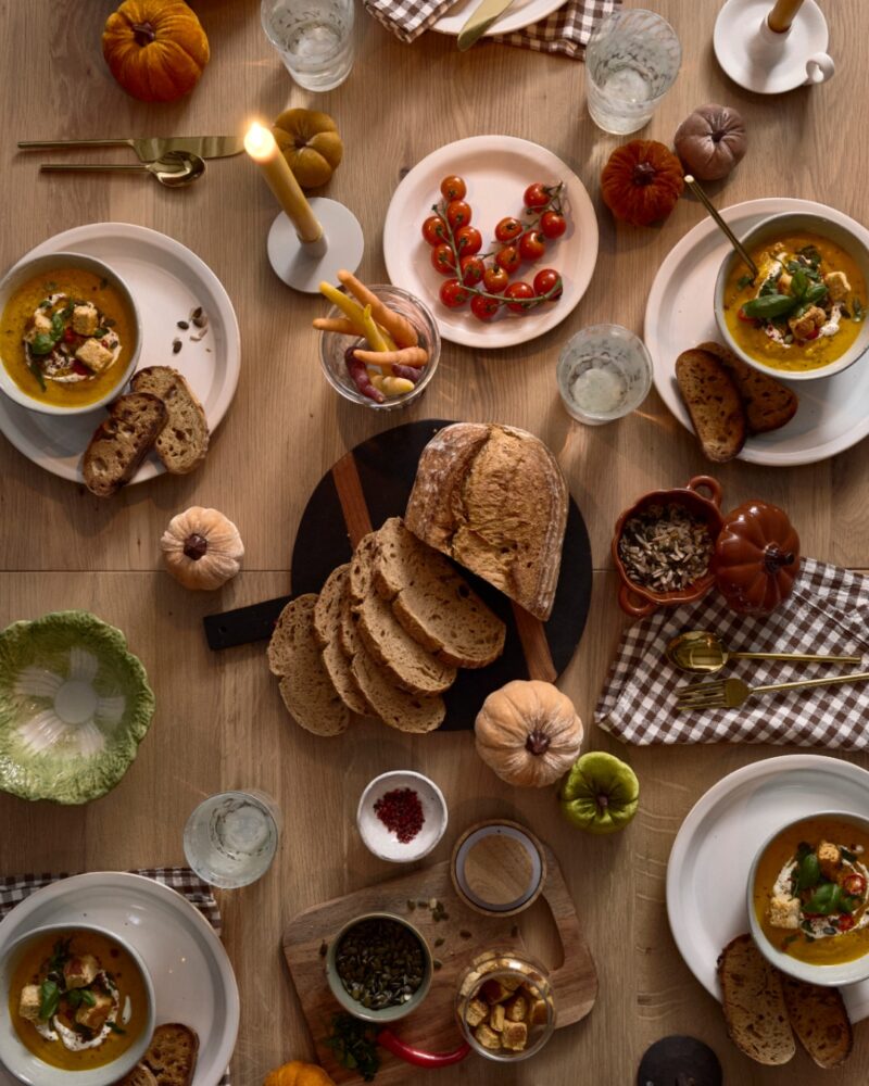 A tabletop-dining room furniture-birds eye table view-natural oak dining table-gingham brown napkins-white crockery-velvet pumpkins-tapered candles-candlelit-soup bowls-chunky bread