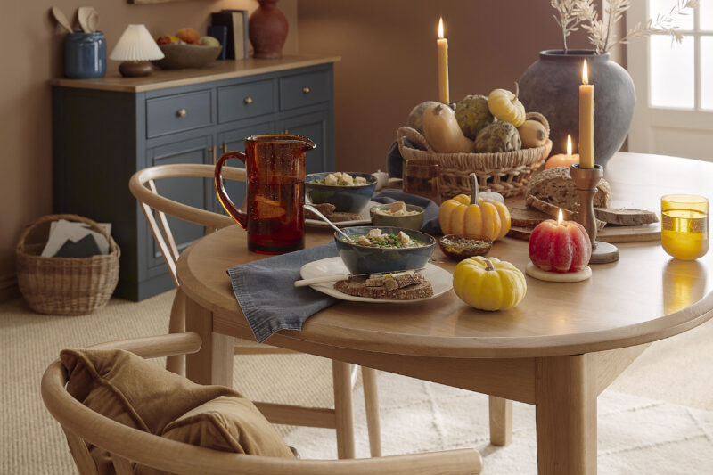A dining table, chairs, and sideboard-dining room furniture-natural oak oval dining table-wishbone style dining chairs-warm brown walls-autumnal tablescape-halloween decor-neutral textured rug-sisal carpet-woven basket filled with autumn vegetables-pumpkins-pumpkin candles-green soup-off white french doors-amber water jug-scatter cushions-dark blue sideboard