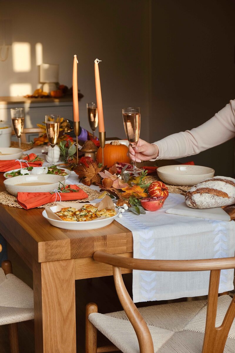 A dining table, chairs, and person-dining room furniture-neutral walls-natural oak dining table-wishbone style dining chairs-neutral walls-person's arm holding glass-neutral table runner-pumpkin centrepiece-autumnal tablescape-halloween decor-orange napkins-off white crockery-rustic bread-sideboard-tapered candles in brass candleholders