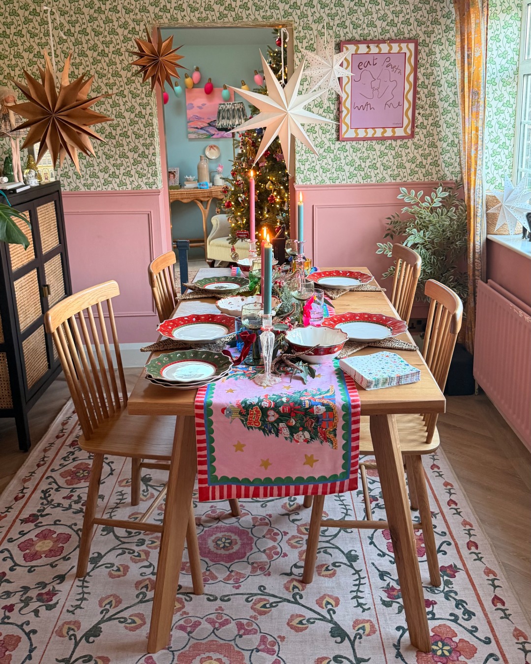 A dining table and chairs-dining room furniture-wooden dining table with play legs-spindle back wooden chairs-floral patterned rug-christmas table runner-pink and green tapered candles-christmas crockery-paper star decorations-pink painted panelling-green patterned wallpaper-black and rattan storage unit
