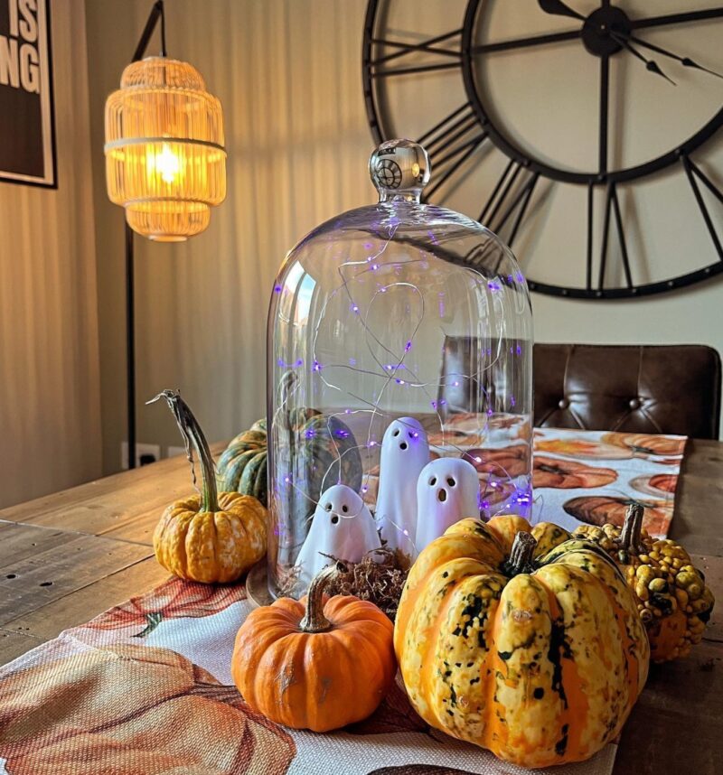 A dining table and wall clock-dining room furniture-wooden dining table-brown leather chair-oversized black wall clock-pumpkin centrepiece-ceramic ghosts under cloche-pumpkin table runner-floor standing rattan lamp