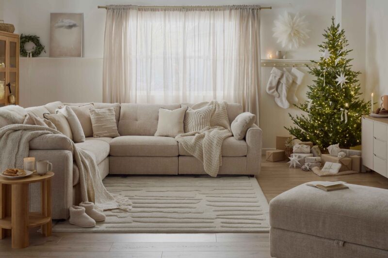A corner sofa, footstool, side table, and sideboard-display cabinet-living room furniture-beige corner modular sofa-beige footstool-tactile scatter cushions-textured neutral rug-knitted throws-warm wooden side table-neutral painted large sideboard-christmas tree-warm wooden display cabinet-fairy lights-off white walls-eucalyptus wreath-neutral stockings-wrapped christmas presents-cosy slippers-candle-mince pies