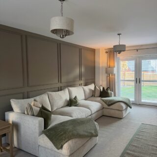 A sofa and side table-living room furniture-beige U-shaped corner chaise sofa-green scatter cushions-green throw-green edged jute rug-beige carpet-brown beaded feature wall-double doors with shutters-floor standing lamp-beige and metal ceiling lights-wooden side table-off white ceiling